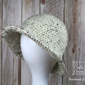 May include: A crocheted hat with a speckled pattern in cream, brown, and black. The hat has a wide brim and is shown on a white mannequin head. The hat is handmade by Tiny Crafty Corner.