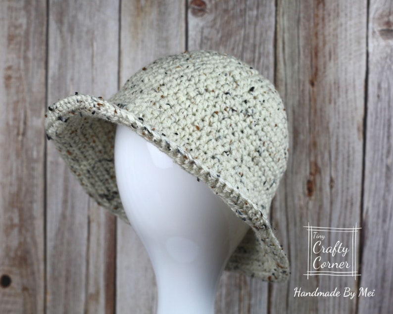 May include: A crocheted hat with a speckled pattern in cream, brown, and black. The hat has a wide brim and is shown on a white mannequin head. The hat is handmade by Tiny Crafty Corner.