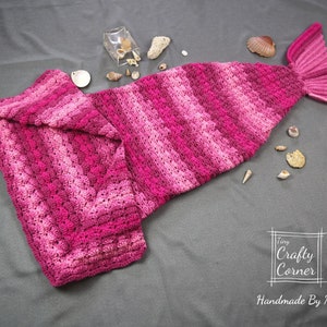 May include: A pink and white striped crocheted mermaid tail blanket. The tail is folded in half and the fin is visible. The blanket is on a gray surface with seashells scattered around it. The text "Tiny Crafty Corner Handmade By Mei" is visible in the lower right corner of the image.