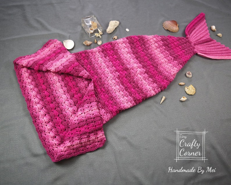 May include: A pink and white striped crocheted mermaid tail blanket. The tail is folded in half and the fin is visible. The blanket is on a gray surface with seashells scattered around it. The text "Tiny Crafty Corner Handmade By Mei" is visible in the lower right corner of the image.