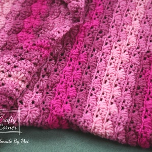 May include: A close-up of a pink and purple crocheted blanket with a textured pattern. The blanket is folded in half and the edges are visible. The text "Tiny Crafty Corner Handmade By Mei" is visible in the lower left corner of the image.