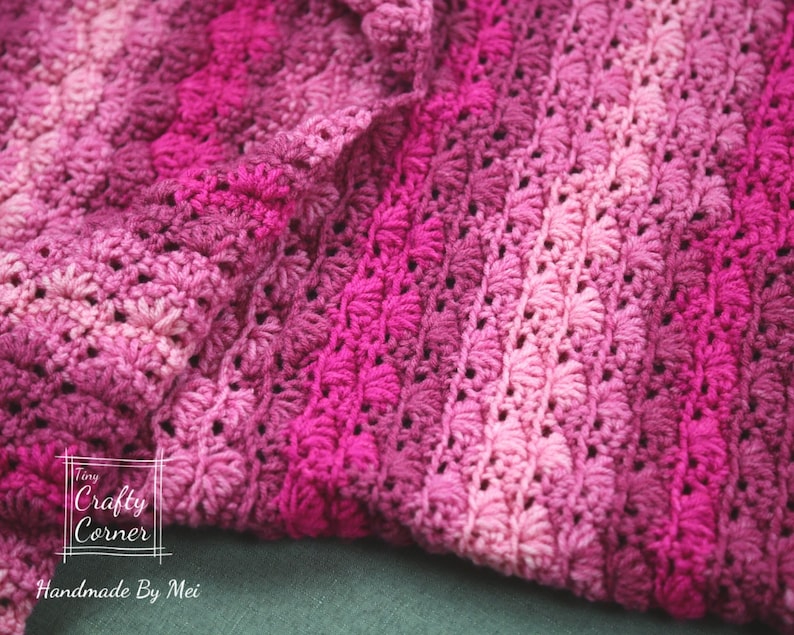 May include: A close-up of a pink and purple crocheted blanket with a textured pattern. The blanket is folded in half and the edges are visible. The text "Tiny Crafty Corner Handmade By Mei" is visible in the lower left corner of the image.