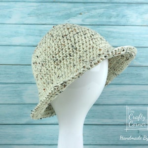 May include: A crocheted hat in a cream color with brown flecks. The hat has a wide brim and is sitting on a white mannequin head. The background is a light blue wooden surface. The text "Tiny Crafty Corner Handmade By Mei" is visible in the bottom right corner of the image.