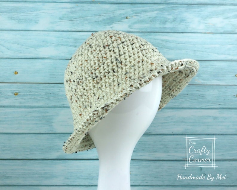 May include: A crocheted hat in a cream color with brown flecks. The hat has a wide brim and is sitting on a white mannequin head. The background is a light blue wooden surface. The text "Tiny Crafty Corner Handmade By Mei" is visible in the bottom right corner of the image.