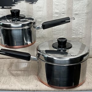 May include: Two stainless steel pots with lids and black handles. The pots have a copper-colored base. One pot is slightly smaller and sits above the other. The lids have black knobs.