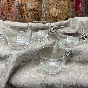 May include: Four clear glass mugs with rounded handles. The mugs are empty and sitting on a light brown fabric surface.