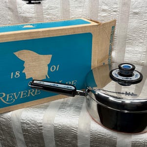 May include: Stainless steel Revere saucepan with a black handle and lid. The lid has a black knob with a blue center. The box is blue and tan with the name "Revere" and the number "1801" printed on it.