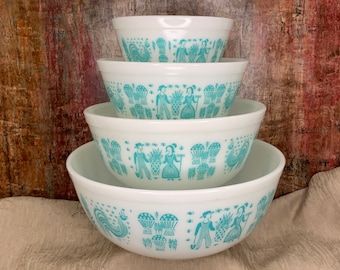 May include: A set of four white Pyrex mixing bowls stacked together. Each bowl features a turquoise design of people, roosters, and baskets of produce. The bowls are nested, with the largest bowl at the bottom and the smallest at the top.