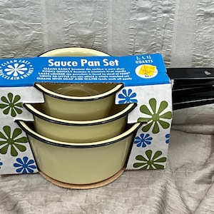 May include: A vintage sauce pan set in its original box. The set includes three pans in varying sizes, with a pale yellow enamel finish and black trim. The box features a blue and green floral design and text that reads "Sauce Pan Set".