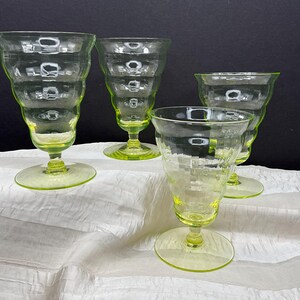 May include: Four vintage, light green-tinted glass goblets with a textured, tiered design. The goblets have a flared rim, a round base, and a stem. The glasses are arranged on a white, textured surface against a dark background.