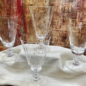 May include: Four clear glass drinking glasses with etched designs on a white fabric surface.