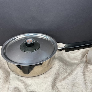May include: Stainless steel saucepan with a lid. The pot has a black handle and a black knob on the lid. The pot is sitting on a beige surface with a dark background.