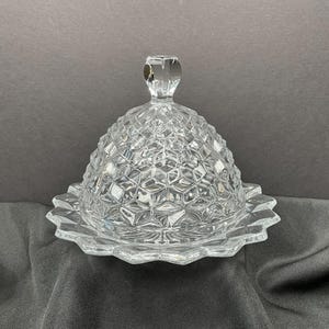 May include: A clear, faceted glass butter dish with a domed lid and a scalloped base. The lid features a geometric pattern and a small, decorative handle. The dish is designed to hold butter and is suitable for kitchen use.