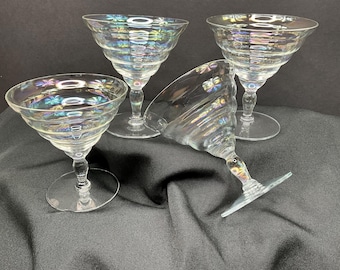 May include: Four clear, iridescent champagne coupe glasses with a spiral design. The glasses have a wide, shallow bowl and a stem with a circular base. The glasses are arranged on a black surface.