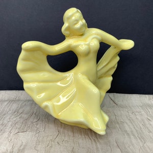 May include: A yellow ceramic figurine of a woman dancing in a flowing dress. The figurine is in a mid-twirl pose with her arms outstretched.