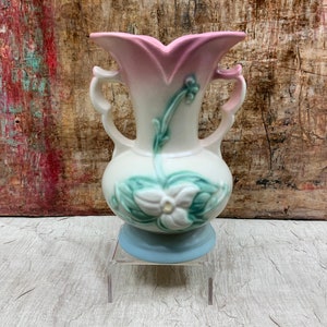 May include: A vintage ceramic vase with a pink and white ombre glaze. The vase features a raised floral design with green leaves and white flowers. The vase has two handles and a blue base.