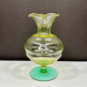 May include: A clear glass vase with a ruffled top and a bulbous body. The vase has a green base and a yellow-green hue throughout. The textured glass catches the light, creating a decorative effect. The vase is approximately 8 inches tall.