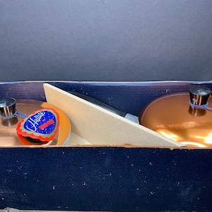 May include: Two copper-colored cookware lids with black handles and tags inside a blue box. The tags read "Hallite" with a red, white, and blue design. A beige divider separates the lids.