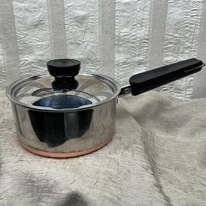 May include: Stainless steel saucepan with a clear glass lid and a black handle. The pot has a copper-colored base and a black knob on the lid. The handle is attached to the side of the pot. The pot is sitting on a light-colored surface.
