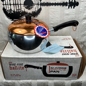 May include: Vintage Hall-ite all-aluminum sauce pan with a copper-colored lid and black handle. The pan is sitting on its original box, which features a graphic of the pan. The box reads "wear-ever hall-ite".
