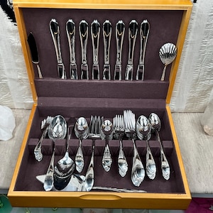 May include: A wooden box with a brown velvet interior containing a set of silver flatware. The flatware includes knives, forks, spoons, and serving pieces. The box is open and the flatware is arranged in a neat and organized manner.