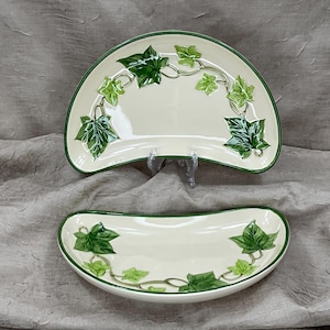 May include: Two off-white ceramic serving dishes with a green ivy leaf design. The dishes have a green trim and are kidney-shaped. The larger dish is propped up on a clear stand. The ivy leaves are in varying shades of green.
