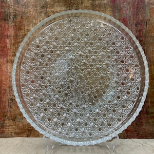 May include: A clear glass serving tray with a decorative pattern of small, raised flowers. The tray has a scalloped edge and is perfect for serving snacks or desserts.
