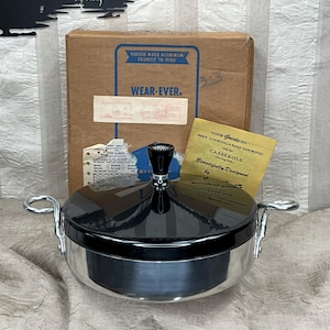 May include: A vintage Wear-Ever casserole dish with a black lid and silver-toned body. The dish has two handles and a black knob on the lid. The original box and a guide to best cooking are behind the dish.