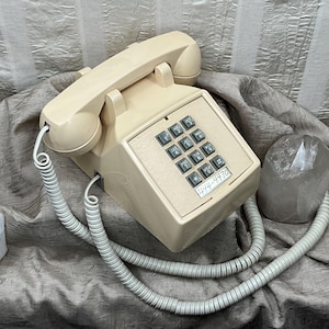 May include: A vintage beige telephone with a keypad and handset, featuring a coiled cord. The phone has a label with the number 444-4470. Two quartz crystals are placed near the phone.