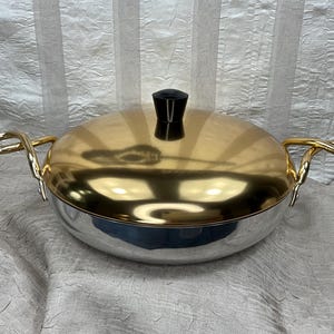 May include: A silver and gold-toned serving dish with a lid and two gold handles. The lid is gold, and the body of the dish is silver. The lid has a black handle. The handles are curved and attached to the sides of the dish.
