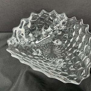 May include: Clear glass serving bowl with a diamond pattern and a scalloped edge. The bowl has a starburst design in the center and sits on three small feet. The bowl is likely used for serving snacks or desserts.