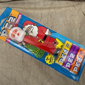 May include: A PEZ candy dispenser featuring a Santa Claus figure in a red suit, packaged in a blue cardboard blister pack with three candy refills in yellow, purple, and orange wrappers. The packaging includes the text PEZ, DISPENSER, and Fun 'n Games On Back.