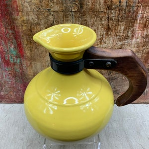 May include: A yellow ceramic pitcher with a wooden handle and a black metal band. The pitcher has a rounded body and a spout. The handle is attached with metal rivets. The pitcher is likely for serving beverages.