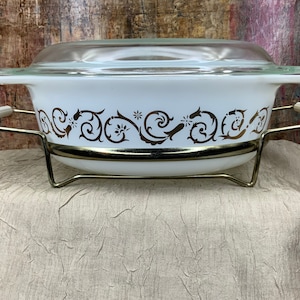 May include: White casserole dish with a brown floral design and a glass lid. The dish is sitting on a metal stand with handles.