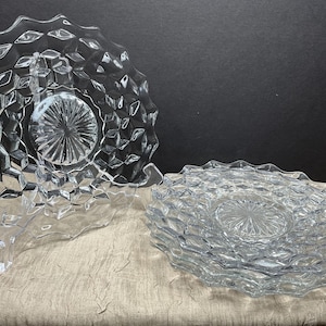 May include: Clear glass plates with a diamond-shaped pattern and scalloped edges. One plate is displayed on a clear stand, while a stack of plates rests on a neutral-colored surface. The plates have a textured, faceted design.