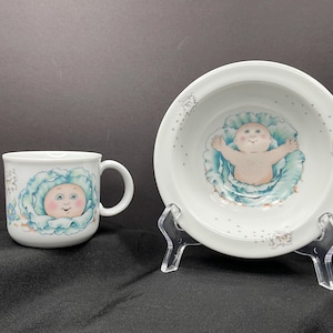 May include: White ceramic mug and bowl set featuring a baby design. The mug and bowl have a baby face surrounded by a blue and green cabbage-like design. The bowl is on a clear plastic stand.