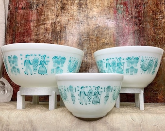 May include: Three white ceramic mixing bowls with a blue and green folk art design. The bowls are nested inside each other and are on a white stand.