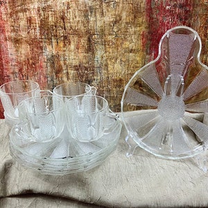 May include: Set of four clear glass teacups and saucers with a textured design. The cups and saucers are stacked on top of each other. A large clear glass plate with a textured design is in the background.