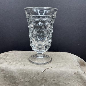 May include: A clear, vintage-style glass with a pedestal base. The glass features a textured, geometric pattern on the body and a smooth rim. The glass is likely used for serving beverages and is displayed on a textured surface.