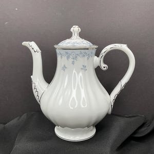 May include: White porcelain teapot with a lid, featuring a delicate blue floral pattern. The teapot has a curved spout and handle, with black trim outlining the edges. A classic piece for serving tea.