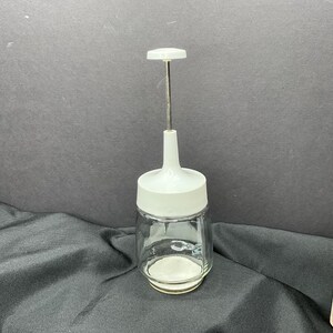 May include: A clear glass jar with a white plastic top and a metal rod extending upwards. The jar has a white base and a white handle at the top. The jar is likely a kitchen tool for chopping or mincing.