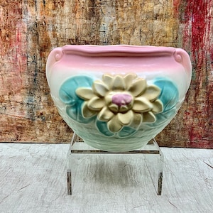 May include: A vintage ceramic planter with a pink, blue, and white glaze. The planter features a raised, white flower with a pink center. The planter is on a clear acrylic stand.