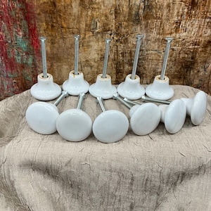 May include: A set of ten white ceramic cabinet knobs, each featuring a smooth, rounded top and a long, threaded metal bolt extending from the base. The knobs are arranged in two rows, with five standing upright and five lying on their sides.