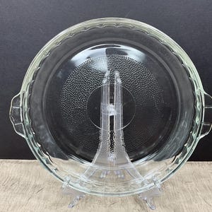 May include: A clear glass pie dish with a scalloped edge and two side handles. The dish sits on a clear plastic stand. The interior of the dish has a textured, dotted pattern. The background is dark.