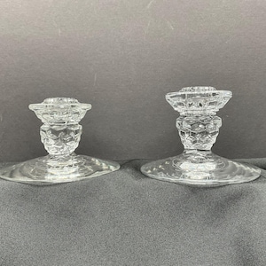 May include: Two clear glass candle holders. Each has a round base, a textured stem, and a circular top designed to hold a candle. The glass has a faceted design, adding visual interest. The candle holders are displayed on a dark surface.