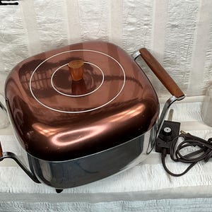 May include: A square electric skillet with a polished copper-colored lid featuring two white concentric circles and a wooden knob. The base is stainless steel with two wooden side handles and a detachable black temperature control cord.