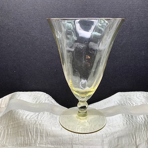 May include: A clear glass goblet with a fluted design and a gold rim. The goblet is sitting on a white fabric surface.