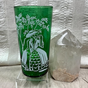 May include: A green glass tumbler with a white silhouette design of a woman in a dress and hat, standing under a tree. Beside it is a clear quartz crystal point.