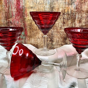 May include: Four red glass cocktail glasses with a fluted design. The glasses are on a white and gray striped surface.