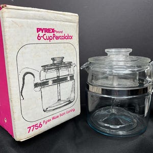May include: A clear glass Pyrex 6-Cup Percolator with a silver band and handle, next to its original box. The box is pink and white with the words "Pyrex brand 6-Cup Percolator" printed on it. The percolator is a vintage kitchen appliance.